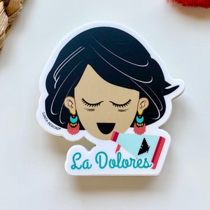 May include: A sticker featuring a cartoon illustration of a woman with black hair and closed eyes. She is wearing turquoise earrings and a red and white patterned dress. She is holding a megaphone with a turquoise and white design. The text "La Dolores" is written in turquoise below the woman.
