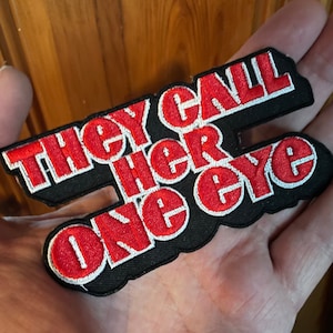 Könnte beinhalten: Schwarzer und roter gestickter Aufnäher mit dem Text "They Call Her One Eye" in einer fetten, blockartigen Schrift.