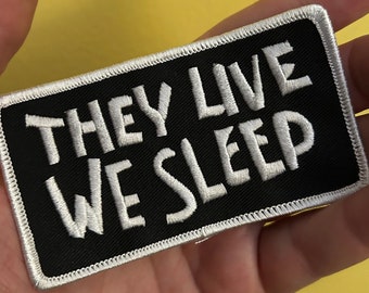 May include: Black embroidered patch with white stitching. The patch reads "They Live We Sleep".