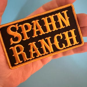 May include: A rectangular black patch with the words "SPAHN RANCH" embroidered in gold thread. The patch has a gold border and is likely designed to be sewn onto clothing or accessories.