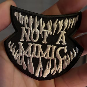 Peut inclure: Patch brodé noir et blanc avec une forme de bouche dentelée et le texte "NOT A MIMIC" en blanc.