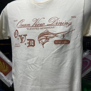 May include: Cream-colored t-shirt featuring a brown graphic with the text "Ocean View Dining" and "Elevated Menswear." The design includes a marlin illustration and the text "Since 1982." The shirt is displayed on a mannequin.
