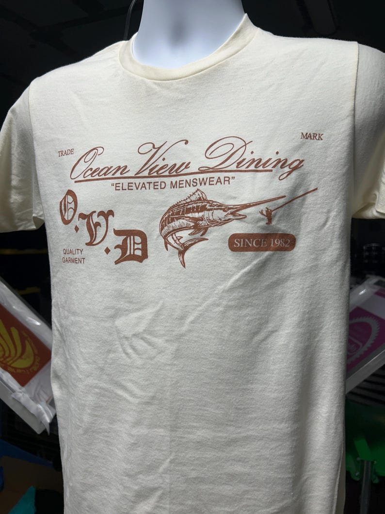 May include: Cream-colored t-shirt featuring a brown graphic with the text "Ocean View Dining" and "Elevated Menswear." The design includes a marlin illustration and the text "Since 1982." The shirt is displayed on a mannequin.