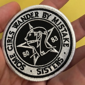 Puede incluir: Un parche bordado en blanco y negro con el texto "Some Girls Wander By Mistake" y "Sisters" alrededor de una estrella de cinco puntas con una figura estilizada en su interior. La estrella tiene los números "80" y "83" en dos de sus puntas.