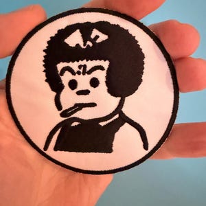 Op de afbeelding: Een ronde patch met een zwart-wit stripfiguur met een afro en een serieuze uitdrukking. Het personage staat centraal op een witte achtergrond met een zwarte rand. De patch wordt in een hand gehouden.