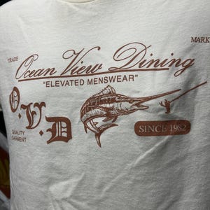 May include: Cream-colored t-shirt with a brown graphic featuring a marlin, a fisherman, and the text "Ocean View Dining" and "Elevated Menswear". The shirt also includes the text "Since 1982".