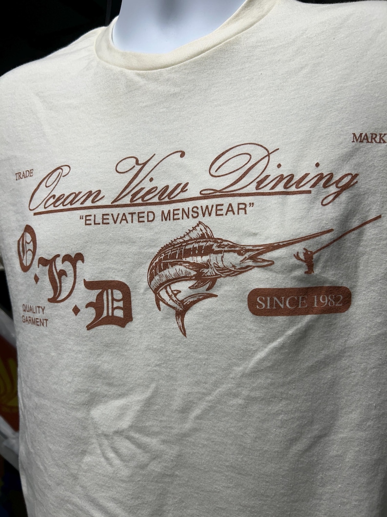 May include: Cream-colored t-shirt with a brown graphic featuring a marlin, a fisherman, and the text "Ocean View Dining" and "Elevated Menswear". The shirt also includes the text "Since 1982".