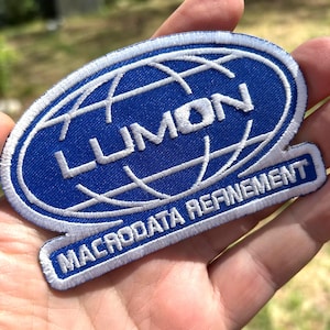 Puede incluir: Un parche bordado azul y blanco con la palabra "LUMON" dentro de un óvalo con un diseño de globo. Debajo, una sección rectangular con las palabras "MACRODATA REFINEMENT".