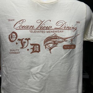 May include: Cream-colored t-shirt with a brown graphic featuring a marlin and the text "Ocean View Dining, Elevated Menswear, Since 1992." The shirt also has the letters "O.V.D." in a decorative font, and the words "Trade" and "Quality Garment."