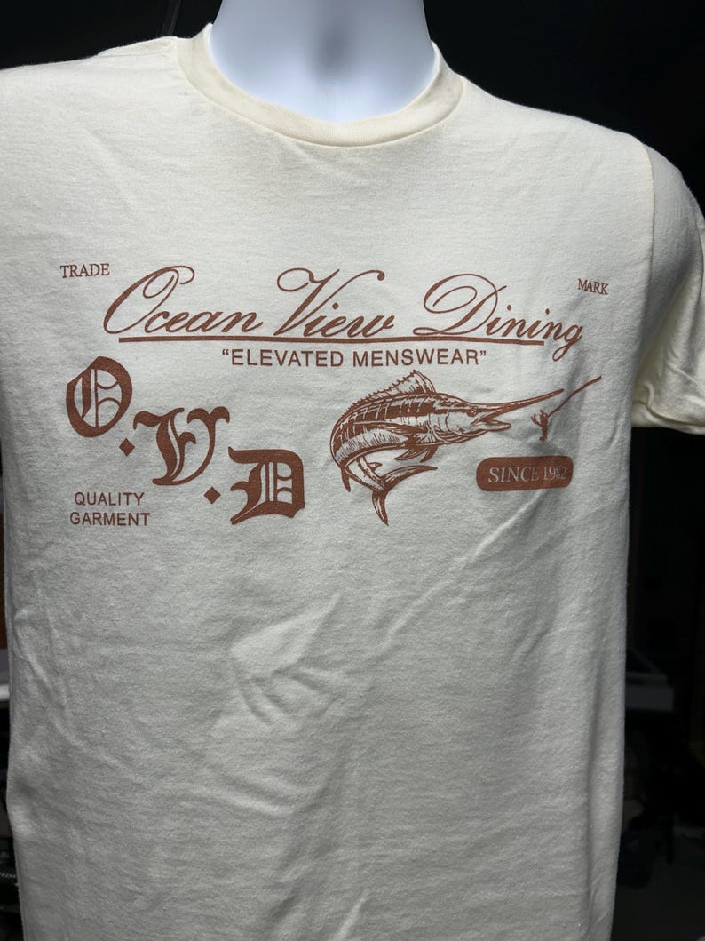 May include: Cream-colored t-shirt with a brown graphic featuring a marlin and the text "Ocean View Dining, Elevated Menswear, Since 1992." The shirt also has the letters "O.V.D." in a decorative font, and the words "Trade" and "Quality Garment."
