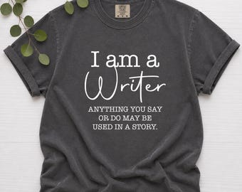 May include: A charcoal gray short-sleeve t-shirt with white text that reads I am a Writer, followed by the sentence ANYTHING YOU SAY OR DO MAY BE USED IN A STORY. The shirt features a crew neckline and a label inside the collar area.