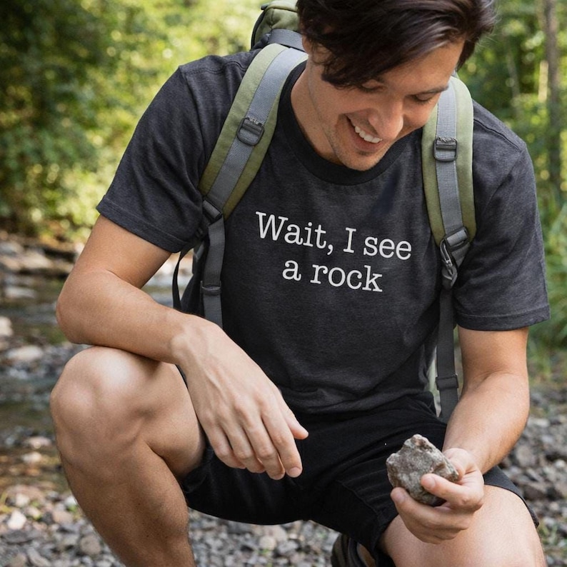 A man in a black t-shirt holding a rock and smiling, with a backpack on, in a forested area.