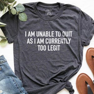 May include: Dark gray t-shirt with the text "I AM UNABLE TO QUIT AS I AM CURRENTLY TOO LEGIT" in white. The shirt is knotted at the bottom and is paired with blue jeans and brown sandals.