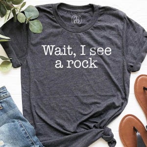 a gray t-shirt with the text "WAIT, I see a rock" printed on it, along with a pair of jeans and brown sandals.
