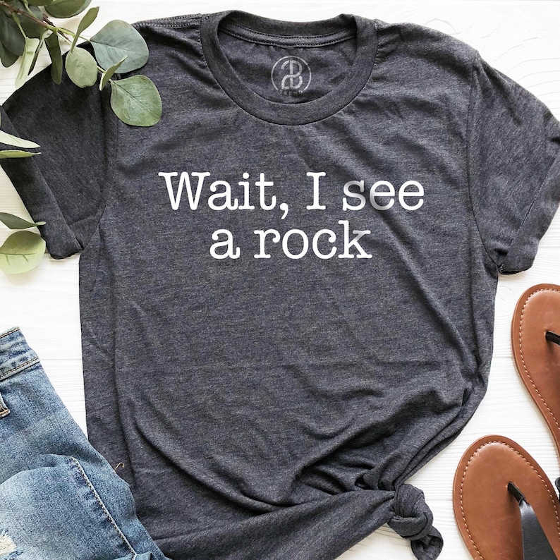 a gray t-shirt with the text "WAIT, I see a rock" printed on it, along with a pair of jeans and brown sandals.