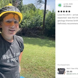 A young boy wearing a black t-shirt with a humorous phrase, "WAIT, I see a rock", standing in a grassy area with trees in the background.