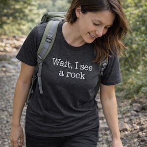A woman wearing a black t-shirt with the text "WAIT, I see a rock" is walking on a dirt path surrounded by greenery.