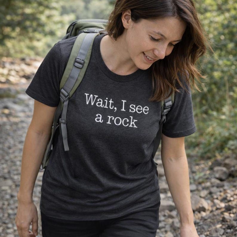 A woman wearing a black t-shirt with the text "WAIT, I see a rock" is walking on a dirt path surrounded by greenery.