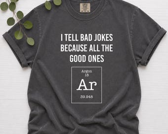 May include: A charcoal gray short-sleeve t-shirt featuring the white text I TELL BAD JOKES BECAUSE ALL THE GOOD ONES followed by a periodic table square for Argon with the symbol Ar, atomic number 18, and atomic weight 39.948 printed in white.