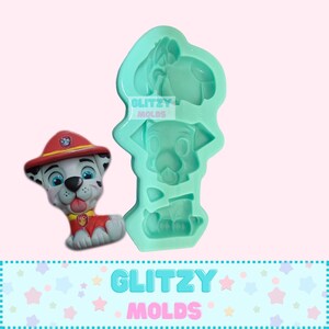May include: A green silicone mold for making a cartoon dog figurine. The mold is shaped like a dog with a red hat and a white and black spotted body. The mold is labeled "Glitzy Molds".