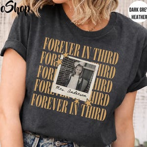 Może przedstawiać: Ciemnoszary melanżowy t-shirt z powtarzającym się złotym napisem "FOREVER IN THIRD" oraz centralną grafiką w stylu polaroida przedstawiającą dziecko z odręcznym napisem "Mrs. Anderson" na dole, ozdobioną dwiema małymi złotymi gwiazdkami.