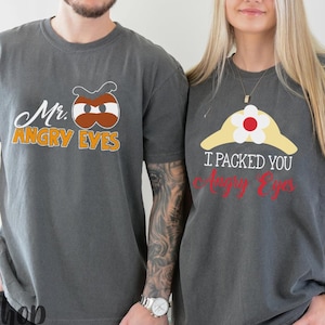 May include: Two grey t-shirts with matching graphics and text. One shirt says "Mr. Angry Eyes" with a cartoon eye design. The other says "I packed you Angry Eyes" with a similar design. The shirts are worn by two people.