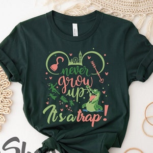 Op de afbeelding: Donkergroen t-shirt met de tekst "never grow up" en "It's a trap!" in een speels lettertype. Het ontwerp bevat een krokodil, een haak en andere Peter Pan-thema graphics. Een casual, comfortabel kledingstuk.
