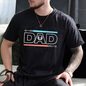 May include: Black t-shirt with a white "DAD" graphic and a blue and red lightsaber design. The shirt has a distressed look and a subtle star pattern.