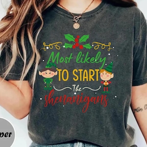 Może przedstawiać: Ciemnoszara koszulka z napisem "Most likely to start the shenanigans" w kolorze zielonym i czerwonym. Koszulka przedstawia dwa kreskówkowe elfy i akcenty ostrokrzewu, idealna na sezon świąteczny.