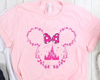 Puede incluir: Una camiseta rosa claro de manga corta con un gráfico de una silueta de orejas de ratón delineada con pequeños lazos rosas de concienciación sobre el cáncer de mama, corazones y huellas de manos. Un lazo rosa con lunares y un castillo rosa.