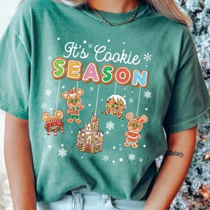May include: Seafoam green t-shirt with the text "It's Cookie Season" in a gingerbread cookie design. The shirt features gingerbread characters and a castle. The shirt is a relaxed fit.