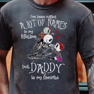 May include: A dark gray t-shirt with a graphic of a skeleton character holding two smaller skeleton characters. The text on the shirt reads "I've been called a lot of names in my lifetime but Daddy is my favorite."