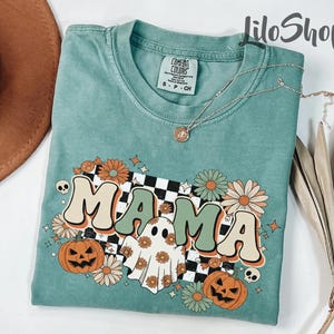 May include: A teal Comfort Colors t-shirt with a retro graphic that says "MAMA". The design includes pumpkins, flowers, a ghost, and a checkered pattern. A silver necklace with a pendant is draped across the shirt.