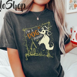 May include: Dark gray t-shirt featuring a graphic of Zero from The Nightmare Before Christmas, with the text "I'M A DOG PERSON". The design is framed with a yellow border. The shirt has short sleeves and a relaxed fit.