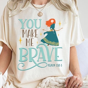 Może przedstawiać: Biały T-shirt z grafiką w kolorze turkusowym i złotym przedstawiającą kobietę z czerwonymi włosami. Grafika zawiera napis "You Make Me Brave" i "Psalm 138:3".