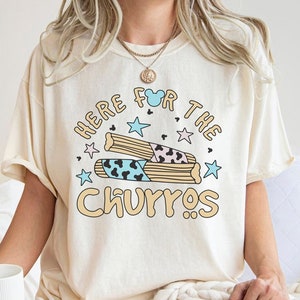 May include: A beige t-shirt featuring the text "Here for the Churros" in a playful font. The text is surrounded by stars and a cartoon illustration of churros.