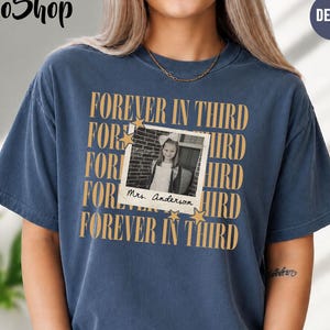 Może przedstawiać: Krótkorękawowa koszulka w kolorze dżinsowego błękitu z powtarzającym się złotym napisem "FOREVER IN THIRD" oraz centralną grafiką w stylu polaroida, zawierającą czarno-białe zdjęcie dziecka z odręcznym podpisem "Mrs. Anderson" poniżej.
