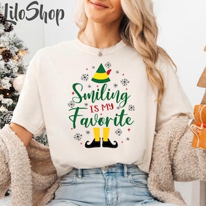 May include: A cream-coloured t-shirt with the text "Smiling is my Favourite" in green script, featuring an elf hat and shoes design. The design includes snowflakes and red accents, perfect for the holiday season.