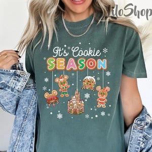 May include: Green t-shirt with the text "It's Cookie Season" in colorful letters. The shirt features gingerbread cookie designs, including a gingerbread castle, gingerbread people, and snowflakes, creating a festive holiday theme.