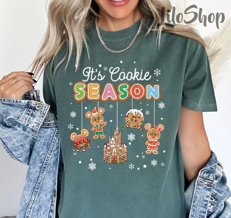 May include: Green t-shirt with the text "It's Cookie Season" in colorful letters. The shirt features gingerbread cookie designs, including a gingerbread castle, gingerbread people, and snowflakes, creating a festive holiday theme.
