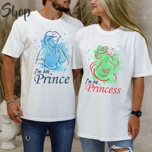 Puede incluir: Dos camisetas blancas con dise&ntilde;os a juego. Una camiseta presenta una ilustraci&oacute;n en acuarela azul de un pr&iacute;ncipe con el texto "I'm her. Prince". La otra camiseta tiene una ilustraci&oacute;n verde y roja de una princesa con el texto "I'm his. Princess."