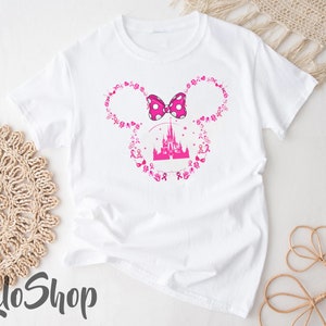 May include: A white short-sleeved t-shirt featuring a pink graphic of a castle silhouette centered within a Mickey Mouse ear outline. The outline is composed of small pink breast cancer awareness ribbons and symbols, topped with a pink polka-dot bow.