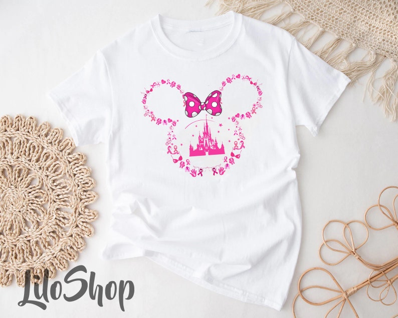 May include: A white short-sleeved t-shirt featuring a pink graphic of a castle silhouette centered within a Mickey Mouse ear outline. The outline is composed of small pink breast cancer awareness ribbons and symbols, topped with a pink polka-dot bow.