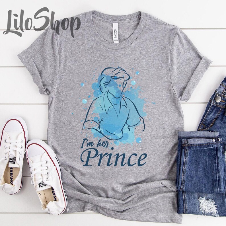 Puede incluir: Una camiseta gris jaspeado con una ilustraci&oacute;n en acuarela azul de un pr&iacute;ncipe y el texto "I'm her Prince". Zapatillas blancas y vaqueros azules son visibles en la imagen.