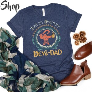 May include: Navy blue t-shirt with the text "Just an ordinary DEMI-DAD" and a cartoon graphic. The shirt is styled with brown leather shoes and a patterned scarf. The Lilo Shop logo is in the top left corner.