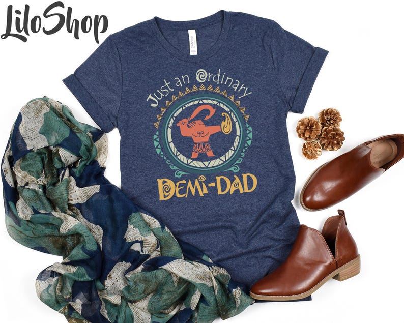 May include: Navy blue t-shirt with the text "Just an ordinary DEMI-DAD" and a cartoon graphic. The shirt is styled with brown leather shoes and a patterned scarf. The Lilo Shop logo is in the top left corner.
