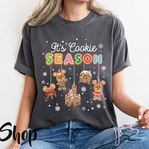 May include: Dark gray t-shirt with the text "It's Cookie SEASON" in colorful letters. The shirt features gingerbread mouse characters and a gingerbread castle. A festive design for the holiday season.