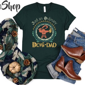May include: Dark green t-shirt with the text "Just an ordinary Demi-Dad" and a graphic of a cartoon figure. The shirt is paired with blue jeans, brown ankle boots, and a patterned scarf with pine cones and a pumpkin.