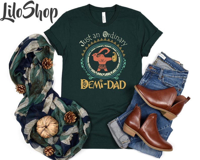 May include: Dark green t-shirt with the text "Just an ordinary Demi-Dad" and a graphic of a cartoon figure. The shirt is paired with blue jeans, brown ankle boots, and a patterned scarf with pine cones and a pumpkin.