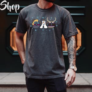 Puede incluir: Una camiseta gris oscuro con un gr&aacute;fico de los personajes de la pel&iacute;cula Frozen. El gr&aacute;fico muestra a Olaf, Elsa y Anna en un dise&ntilde;o estilizado. El texto "DAD" se deletrea usando las cabezas de los personajes.